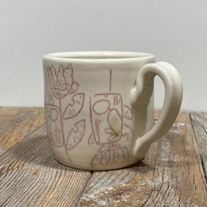 Puede incluir: Taza de cerámica color crema con asa redondeada. La taza presenta un diseño dibujado a mano en un rosa rojizo apagado, que representa flores estilizadas y formas geométricas. La taza está sobre una superficie de madera envejecida.