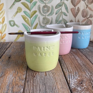 Puede incluir: Tres tazas de cerámica para agua de pintura en colores pastel: verde claro, rosa y azul. Cada taza tiene un borde blanco y las palabras "PAINT WATER" en relieve en la parte delantera. Pinceles descansan en los bordes. Las tazas están sobre una superficie de madera envejecida.