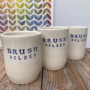 May include: Three off-white ceramic brush holders with the words "BRUSH HOLDER" in navy blue lettering. The holders are cylindrical and sit on a wooden surface. A colorful abstract painting is in the background.