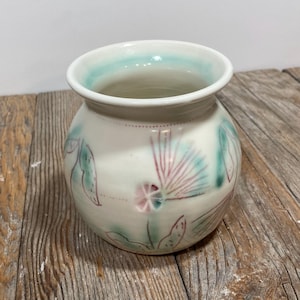 Può includere: Un vaso di ceramica bianco con corpo arrotondato e bordo leggermente svasato. Il vaso è decorato con disegni botanici dipinti a mano nei toni del verde acqua, rosa e verde. Il bordo ha un accento verde acqua. Il vaso è su una superficie di legno.