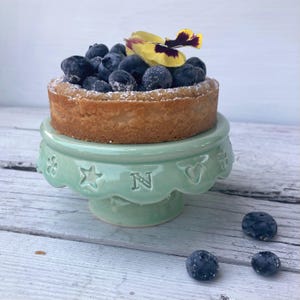 Op de afbeelding: Een kleine, ronde taart met verse bosbessen en een geel-paarse bloem staat op een lichtgroene keramische taartstandaard. De standaard heeft decoratieve uitsparingen van sterren, harten en de letter "N".