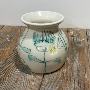 Op de afbeelding: Een kleine, gebroken witte keramische vaas met een afgeronde body en een uitlopende rand. De vaas is versierd met handgeschilderde bloemmotieven in teal, geel en groen. De vaas staat op een verweerd houten oppervlak.