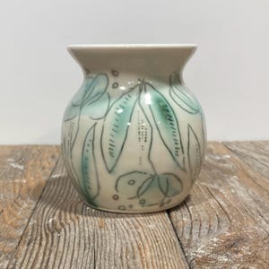 May include: A small, off-white ceramic vase with a rounded body and a slightly flared rim. The vase is decorated with hand-painted teal-colored leaf and dot designs, outlined in black. The vase sits on a weathered wooden surface.