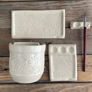 May include: A collection of off-white ceramic art supplies, including a rectangular tray, a brush holder, a paint palette with three wells, and a cylindrical container. All pieces feature a floral relief design and the letter "C". A paintbrush with a red handle is also included.