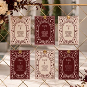 Puede incluir: Un expositor de rejilla metálica dorada con seis tarjetas rectangulares de número de mesa dispuestas en dos filas. Las tarjetas alternan fondos crema y burdeos oscuro con bordes florales ornamentados. Arreglos florales en burdeos y crema.