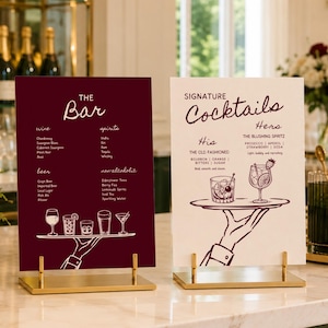 Könnte beinhalten: Zwei rechteckige Menüschilder auf goldenen Ständern. Das linke Schild ist dunkelrot mit weißem Text, der THE Bar liest und Wein, Spirituosen, Bier und alkoholfreie Getränke auflistet. Das rechte Schild ist weiß mit dunkelrotem Text, der SIGNATURE Cocktails liest, und listet His und Her Getränke mit Illustrationen einer Hand auf, die ein Tablett mit Gläsern hält.