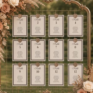 May include: A gold metal frame seating chart display with a grid pattern, featuring twelve white cards labeled Table 1 through 12, each secured with a wax seal. The frame is decorated with arrangements of cream and peach roses and dried pampas grass.