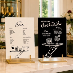 Peut inclure: Deux panneaux de menu rectangulaires sur des supports dorés. Le panneau de gauche est blanc avec du texte noir indiquant THE Bar et liste des vins, spiritueux, bières et boissons sans alcool avec une illustration de main. Le panneau de droite est noir avec du texte blanc indiquant SIGNATURE Cocktails et liste des boissons His et Her avec une illustration de main.