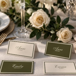 Puede incluir: Cuatro tarjetas de sitio con nombres, en un entorno de boda. Dos tarjetas son de color verde oliva con texto blanco y dos son de color blanquecino con texto negro. Las tarjetas están enmarcadas con un borde fino. Rosas blancas y vegetación en el fondo.