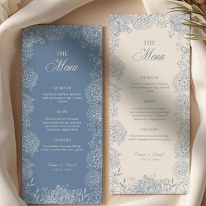 May include: Two wedding menu cards with floral designs. One card is light blue, the other is cream. Both cards have the text "The Menu" at the top, with a list of courses below. The cards are surrounded by flowers and fabric.