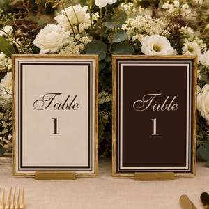 May include: Two rectangular table number signs with gold frames, one featuring a white background with black text and the other a black background with white text, both displaying the script word Table and the number 1 in a serif font.