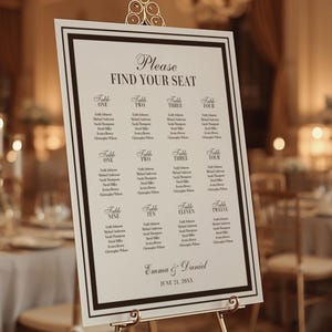 May include: A wedding seating chart on a gold easel. The white sign has a black border and features the words "Please Find Your Seat" at the top. Below are the table assignments and guest names. The names Emma & Daniel and the date are at the bottom.