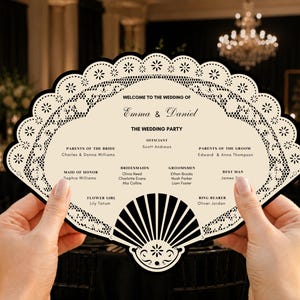 Può includere: Un programma di nozze a forma di ventaglio stampato su cartoncino color crema con un bordo a motivo pizzo nero. Il testo recita Welcome to the wedding of Emma & Daniel, elencando gli sposi, i genitori e il corteo nuziale in inchiostro nero.