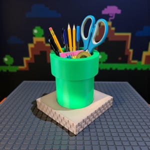 May include: A green pipe-shaped desk tidy, holding pens, pencils, and scissors. It rests on a beige, textured base. The background shows a pixelated video game scene.
