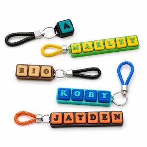 Puede incluir: Cinco llaveros con bases rectangulares de colores y letras en relieve que forman A, MARLEY, RIO, KOBY y JAYDEN. Cada llavero tiene un lazo trenzado en negro, amarillo, negro, azul o naranja, unido mediante un anillo de metal plateado.