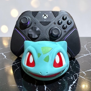 May include: A teal 3D-printed Bulbasaur head sits below a black Xbox controller with purple accents. The Bulbasaur has red and white eyes and a green patch on its head. The controller has the Xbox logo and various buttons.
