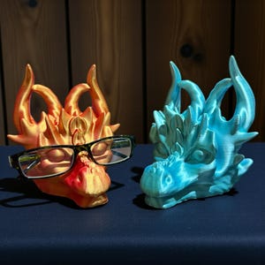 May include: Two dragon head sculptures, one orange and one blue. The orange dragon wears black-framed glasses. The sculptures have detailed features, including horns and textured scales. The dragon heads are displayed on a dark blue surface.