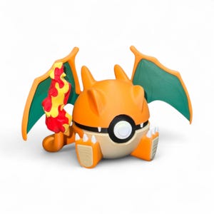 May include: A stylized orange and tan Charizard figure, with green wings and a flame. The figure is in a seated position, with a black and white circle in the center. The figure is a Pokemon.