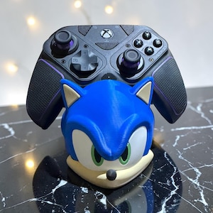 Puede incluir: Un soporte para mando con forma de cabeza de Sonic el Erizo, azul y blanco, que sostiene un mando Xbox negro con detalles morados. El soporte presenta ojos verdes y una nariz negra. El mando tiene varios botones y una cruceta.