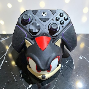 Puede incluir: Mando Xbox negro con detalles morados, apoyado en un soporte para mando de Sonic the Hedgehog negro y rojo. El soporte presenta la cara de Sonic con detalles rojos, blancos y beige. El mando tiene un agarre texturizado.