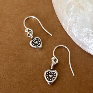 May include: A pair of silver drop earrings featuring small heart-shaped charms. Each charm has a raised central heart surrounded by a dotted border, suspended from a simple silver hook wire. The metal has a polished finish with darkened accents.