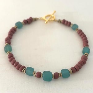 May include: A handmade bracelet featuring a toggle clasp. The bracelet is composed of small, round, reddish-brown beads interspersed with larger, square, teal-colored beads. Gold-colored metal accents add a touch of elegance to the design.
