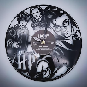 Könnte beinhalten: Eine schwarze Vinyl-Schallplattenuhr mit ausgeschnittenen Silhouetten von Charakteren. Das Zifferblatt enthält die Buchstaben "HP" und den Text "EBONY RECORDS" auf dem Etikett. Die Uhrzeiger sind goldfarben.