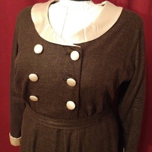 K&ouml;nnte beinhalten: Braunes Vintage-Kleid mit cremefarbenem Kragen und Manschetten. Das Kleid hat eine doppelreihige Front mit gro&szlig;en wei&szlig;en Kn&ouml;pfen. Das Kleid wird auf einer Schaufensterpuppe pr&auml;sentiert.