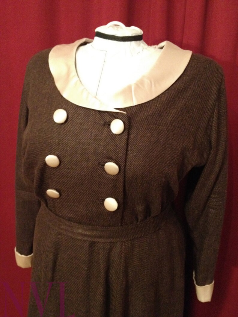 K&ouml;nnte beinhalten: Braunes Vintage-Kleid mit cremefarbenem Kragen und Manschetten. Das Kleid hat eine doppelreihige Front mit gro&szlig;en wei&szlig;en Kn&ouml;pfen. Das Kleid wird auf einer Schaufensterpuppe pr&auml;sentiert.