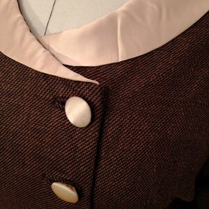 K&ouml;nnte beinhalten: Nahaufnahme einer braunen Tweedjacke mit cremefarbenem Satin-Kragen und drei gro&szlig;en, perlmuttfarbenen Kn&ouml;pfen. Die Jacke hat ein klassisches Design mit taillierter Passform und strukturiertem Stoff.