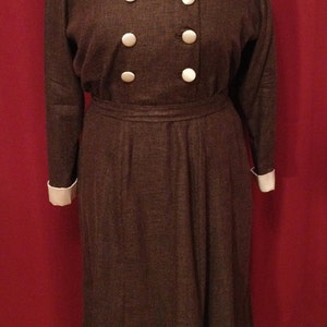 K&ouml;nnte beinhalten: Braunes Vintage-Kleid mit doppelreihiger Front mit sechs gro&szlig;en, runden, wei&szlig;en Kn&ouml;pfen. Das Kleid hat einen cremefarbenen Kragen und Manschetten sowie einen passenden G&uuml;rtel. Das Kleid wird auf einer Schaufensterpuppe pr&auml;sentiert.