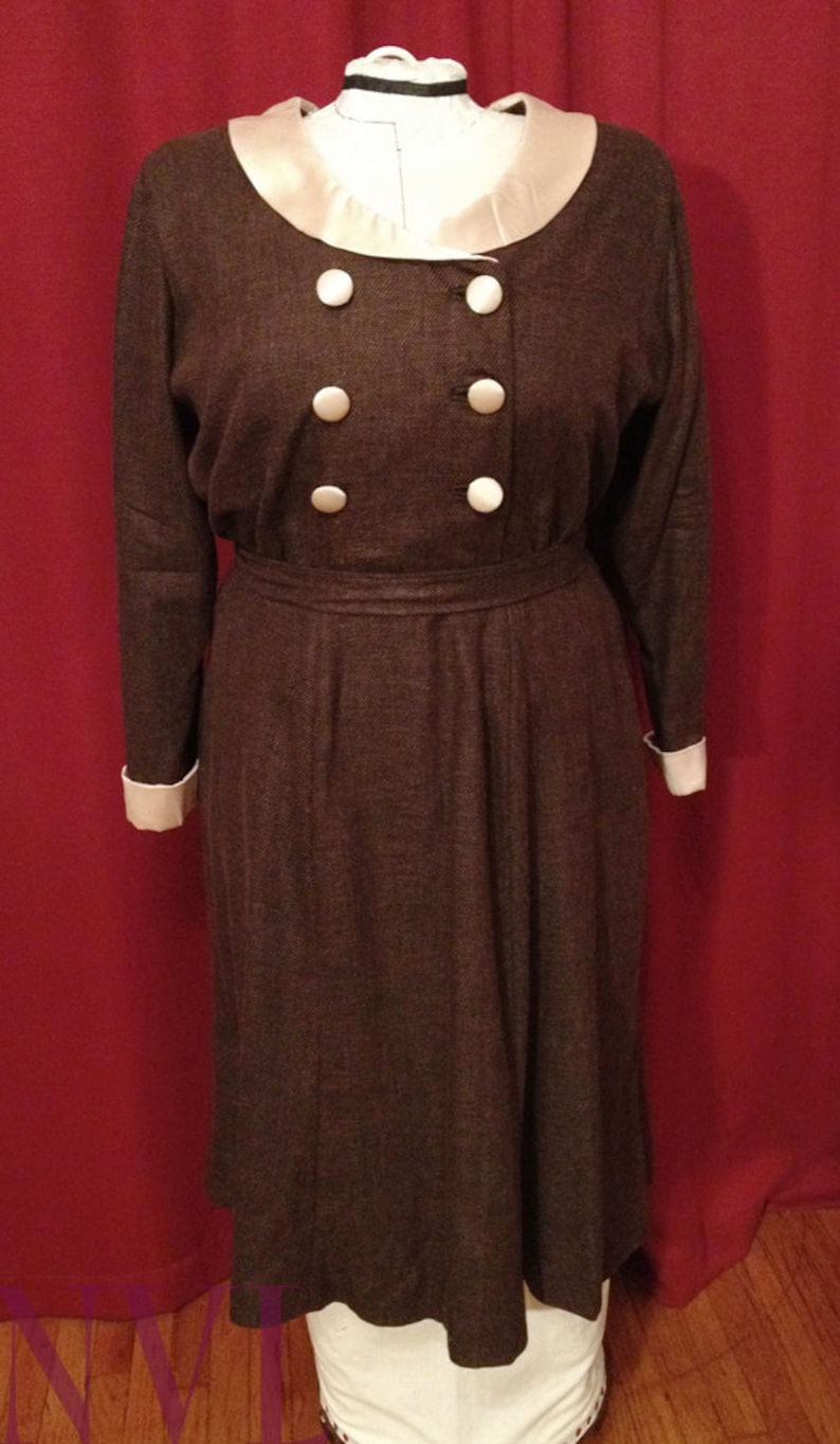 K&ouml;nnte beinhalten: Braunes Vintage-Kleid mit doppelreihiger Front mit sechs gro&szlig;en, runden, wei&szlig;en Kn&ouml;pfen. Das Kleid hat einen cremefarbenen Kragen und Manschetten sowie einen passenden G&uuml;rtel. Das Kleid wird auf einer Schaufensterpuppe pr&auml;sentiert.