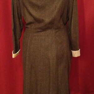 K&ouml;nnte beinhalten: Ein braunes Langarmkleid mit einem Rundhalsausschnitt und wei&szlig;en Manschetten. Das Kleid hat eine taillierte Taille und einen ausgestellten Rock. Das Kleid ist aus einem strukturierten Stoff gefertigt und wird auf einer Schaufensterpuppe pr&auml;sentiert.