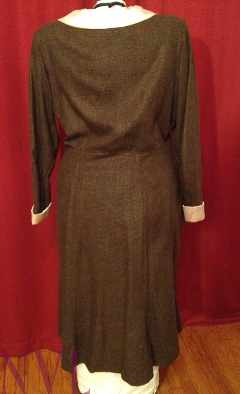 K&ouml;nnte beinhalten: Ein braunes Langarmkleid mit einem Rundhalsausschnitt und wei&szlig;en Manschetten. Das Kleid hat eine taillierte Taille und einen ausgestellten Rock. Das Kleid ist aus einem strukturierten Stoff gefertigt und wird auf einer Schaufensterpuppe pr&auml;sentiert.