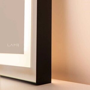 K&ouml;nnte beinhalten: Nahaufnahme eines modernen Spiegels mit schwarzem Rahmen, wei&szlig;em Innenrand und Glasoberfl&auml;che. Das Wort "LAMR" ist dezent in der oberen linken Ecke aufgedruckt. Der Spiegel befindet sich vor einem neutralen Hintergrund.