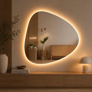 Könnte beinhalten: Ein moderner, organisch geformter Spiegel mit warmem LED-Lichtrand. Der Spiegel hängt an einer neutralen Wand über einer Holzkonsole. Eine Lampe und dekorative Gegenstände ergänzen die minimalistische Einrichtung. Das Wort "LAMR" ist sichtbar.