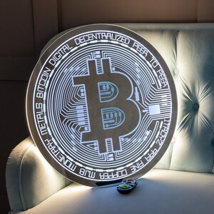Könnte beinhalten: Ein rundes, silberfarbenes, beleuchtetes Schild mit dem Bitcoin-Logo und dem Text "METALS BITCOIN DIGITAL DECENTRALIZED PEER TO PEER FINE COPPER MLB"