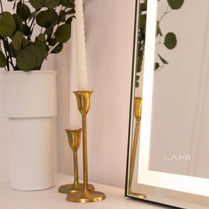 K&ouml;nnte beinhalten: Eine dekorative Anordnung mit einer wei&szlig;en Vase mit gr&uuml;nem Laub, zwei goldenen Kerzenhaltern mit wei&szlig;en Kerzen und einem gerahmten Spiegel. Der Spiegel hat den Text "LAMR" in der unteren rechten Ecke.