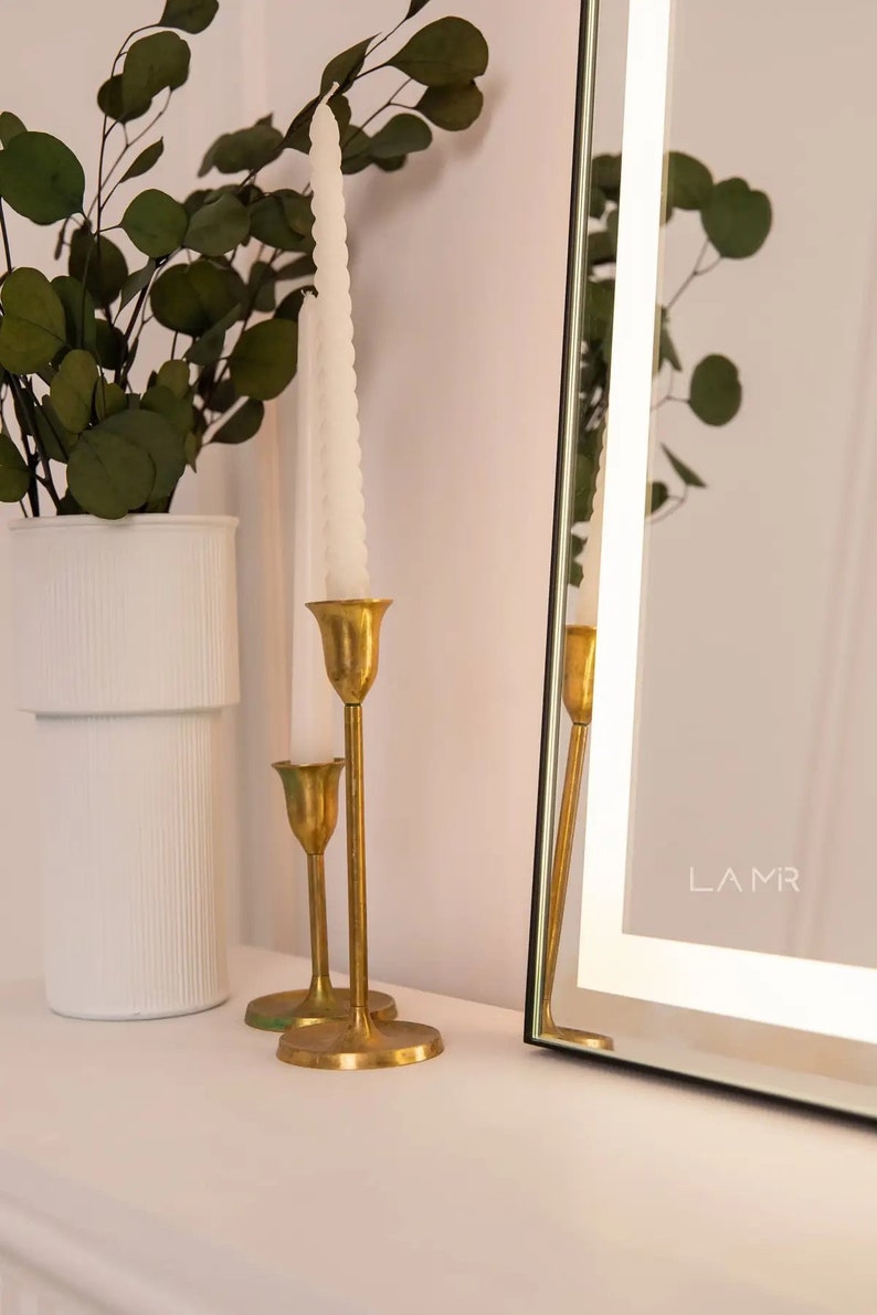 K&ouml;nnte beinhalten: Eine dekorative Anordnung mit einer wei&szlig;en Vase mit gr&uuml;nem Laub, zwei goldenen Kerzenhaltern mit wei&szlig;en Kerzen und einem gerahmten Spiegel. Der Spiegel hat den Text "LAMR" in der unteren rechten Ecke.