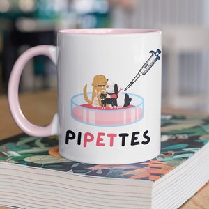 May include: White ceramic mug with a pink rim and handle. The mug features a cartoon illustration of a dog, cat, and rabbit in a petri dish with a pipette. The text "PIPETTES" is printed in pink and black below the illustration.