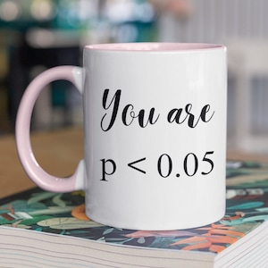 Puede incluir: Una taza de café blanca con un borde y un asa de color rosa. La taza tiene un texto negro que dice "You are p < 0.05".