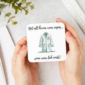 Peut inclure: Sous-verre blanc avec du texte noir qui dit "Tous les héros ne portent pas de capes... certains portent des blouses de laboratoire !" et une illustration de dessin animé d'une blouse de laboratoire blanche avec un stylo dans la poche.