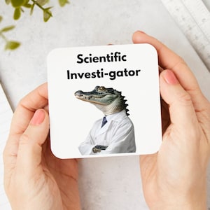 May include: A white coaster with the text "Scientific Investi-gator" in black font. An alligator wearing a white lab coat is pictured on the coaster.