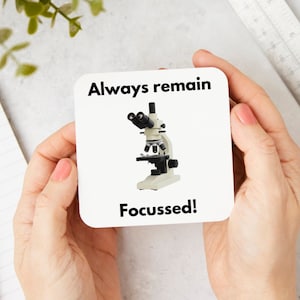 May include: A square white coaster with the text "Always remain Focussed!" and a graphic of a microscope. The microscope is off-white and black. The coaster is held by a person with pink nail polish.