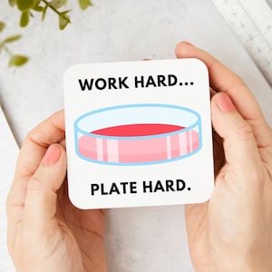 Peut inclure: Sous-verre carré blanc avec du texte noir qui dit "TRAVAILLER DUR... PLAQUER DUR." Un plat de Petri dessiné avec un milieu d'agar rose est au centre du sous-verre.