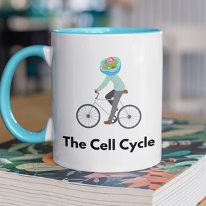 Peut inclure: Mug en céramique blanche avec intérieur et anse bleu clair. L'illustration représente un personnage à vélo avec une tête en forme de cellule. Le texte "The Cell Cycle" est imprimé en dessous.
