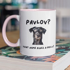 Könnte beinhalten: Weiße Keramiktasse mit rosafarbenem Rand. Die Tasse zeigt einen schwarzen Hund mit Brille und den Text "Pawlow? Der Name sagt dir etwas!"
