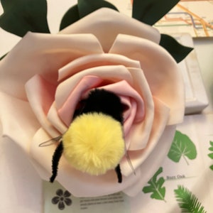 May include: A large, artificial rose in shades of pink and cream, with green leaves. A plush bumblebee with yellow and black fur is nestled inside the rose. The image is set against a background with botanical illustrations.