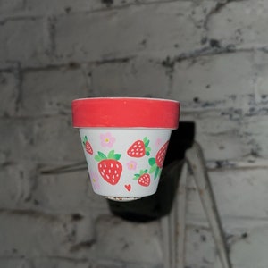 Peut inclure: Un petit pot de fleurs en céramique blanche avec un bord rouge. Le pot est décoré d'un motif peint à la main de fraises et de fleurs roses.