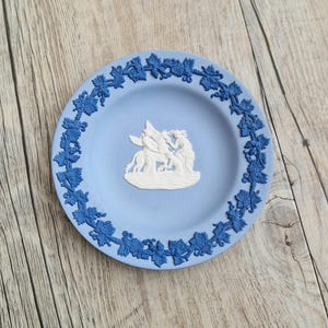May include: A light blue decorative plate with a raised relief design. The plate features a border of blue leaves and a central white relief scene of figures. The plate is set on a wooden surface.